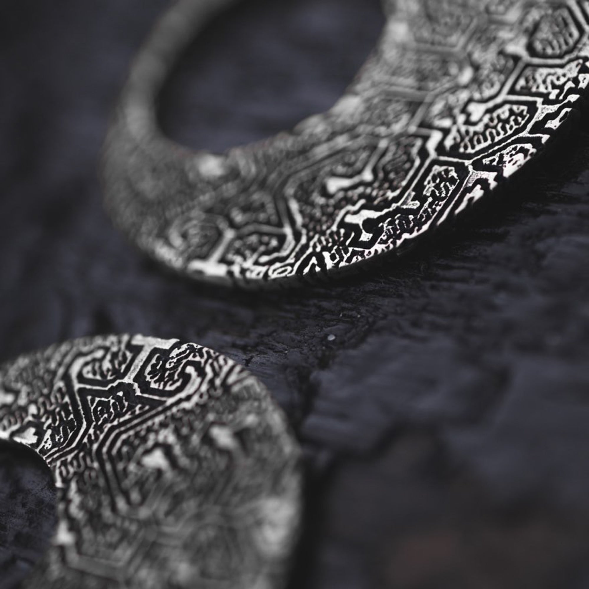 Close-up image of intricately designed OLIVIA Tribal Shipibo Seamless Disc Hoop Earrings in Silver by Kaya Movement, placed on a textured dark surface. The focus is on the detailed patterns etched into these 12 gauge earrings, which feature complex geometric and tribal motifs, adding a sense of elegance to the jewelry.